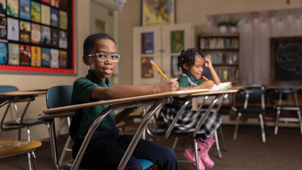 Boy in glasses writing at classroom desk, girl studying behind him, school choice
